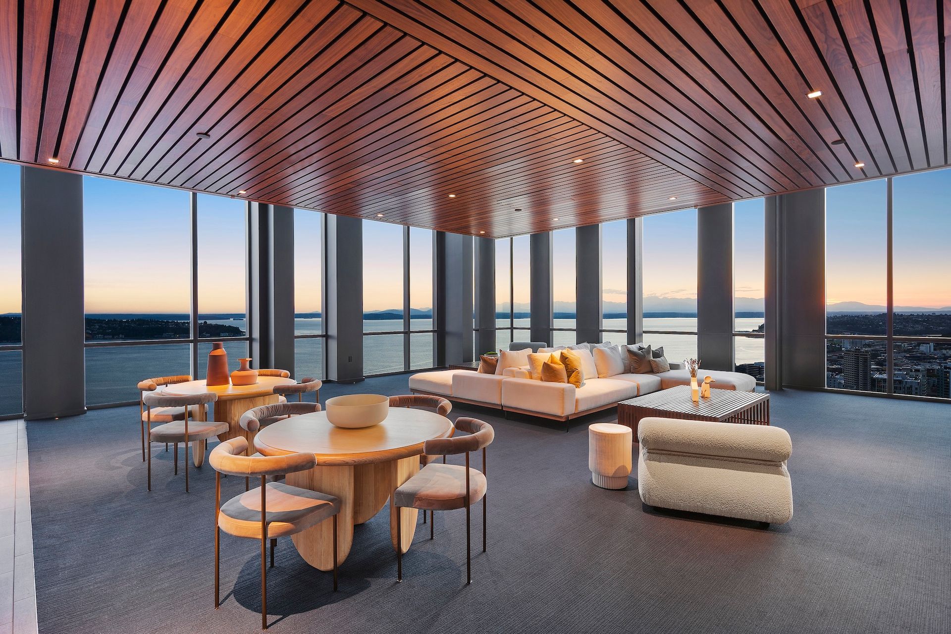Modern lounge with curved wood ceiling, sofas, and tables overlooking a sunset city skyline