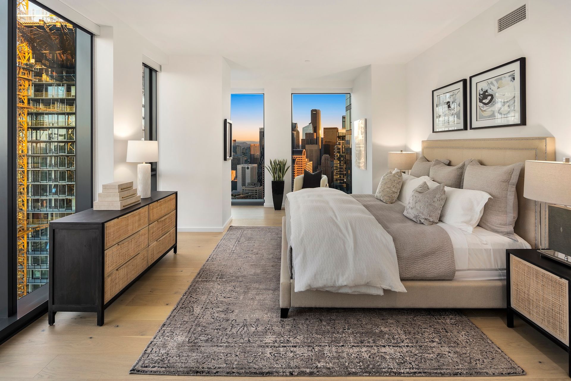 Modern bedroom with a beige bed, gray rug, city skyline view, and black dresser beside floor-to-ceiling windows