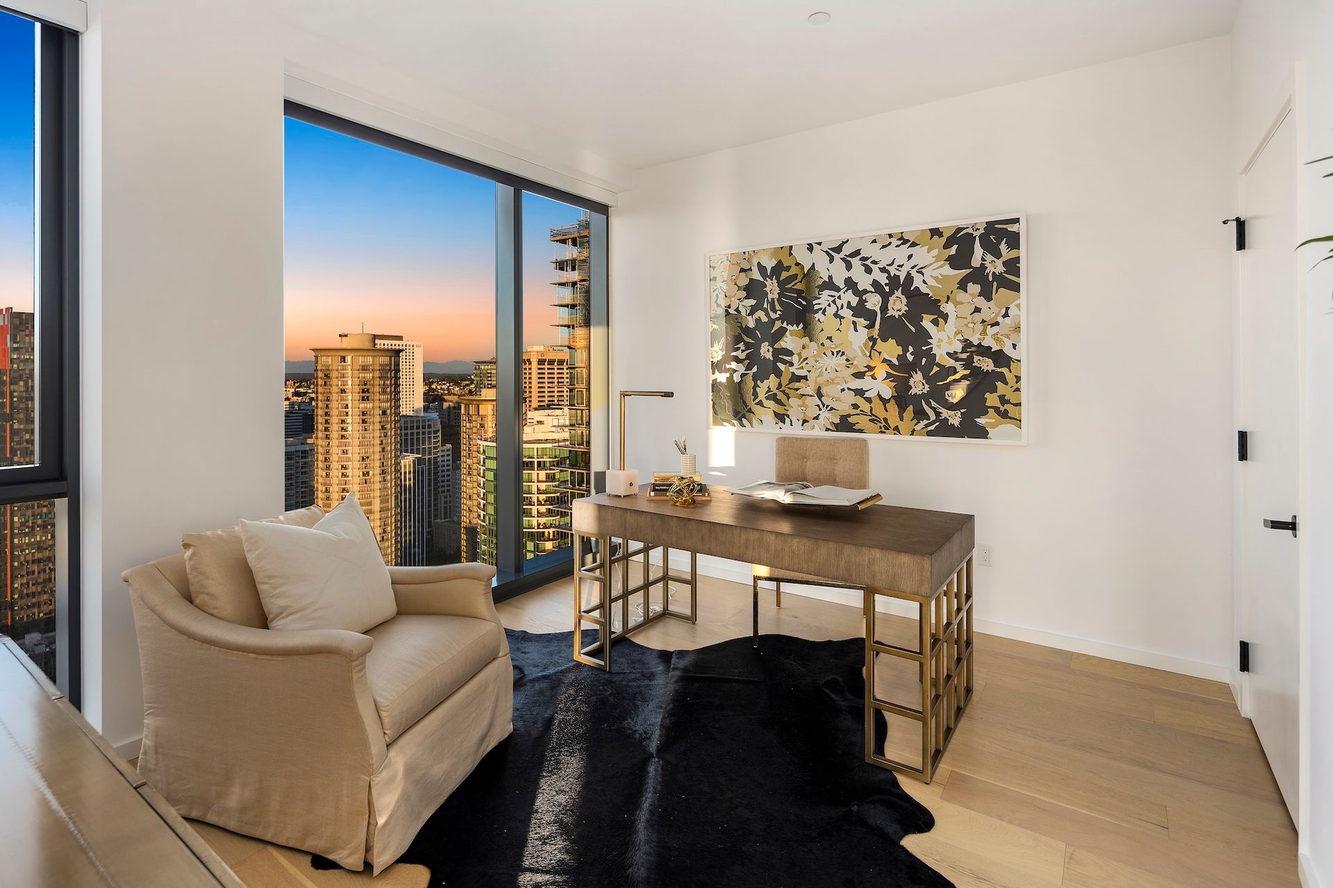 Modern living room with beige armchair, desk, abstract art, and floor-to-ceiling window overlooking city skyline