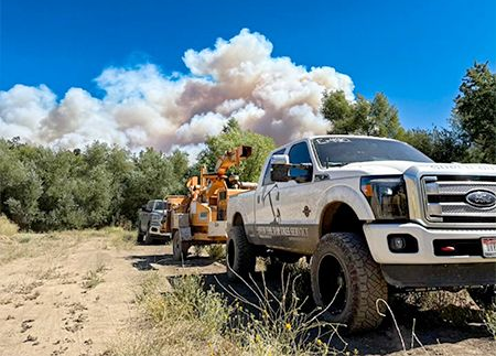White truck with a wood chipper on a dirt road, large wildfire in background.