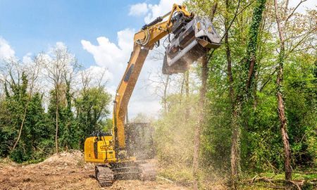 Yellow excavator mulching trees, creating wood chips, on a sunny day.