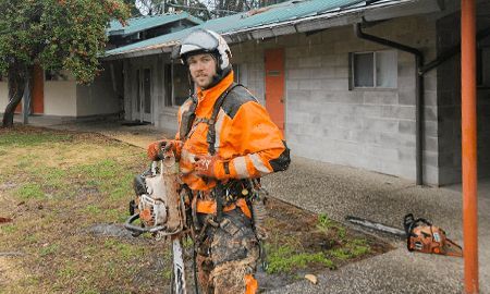 Arborist in orange safety gear, helmet, and harness, holding a chainsaw near a building.