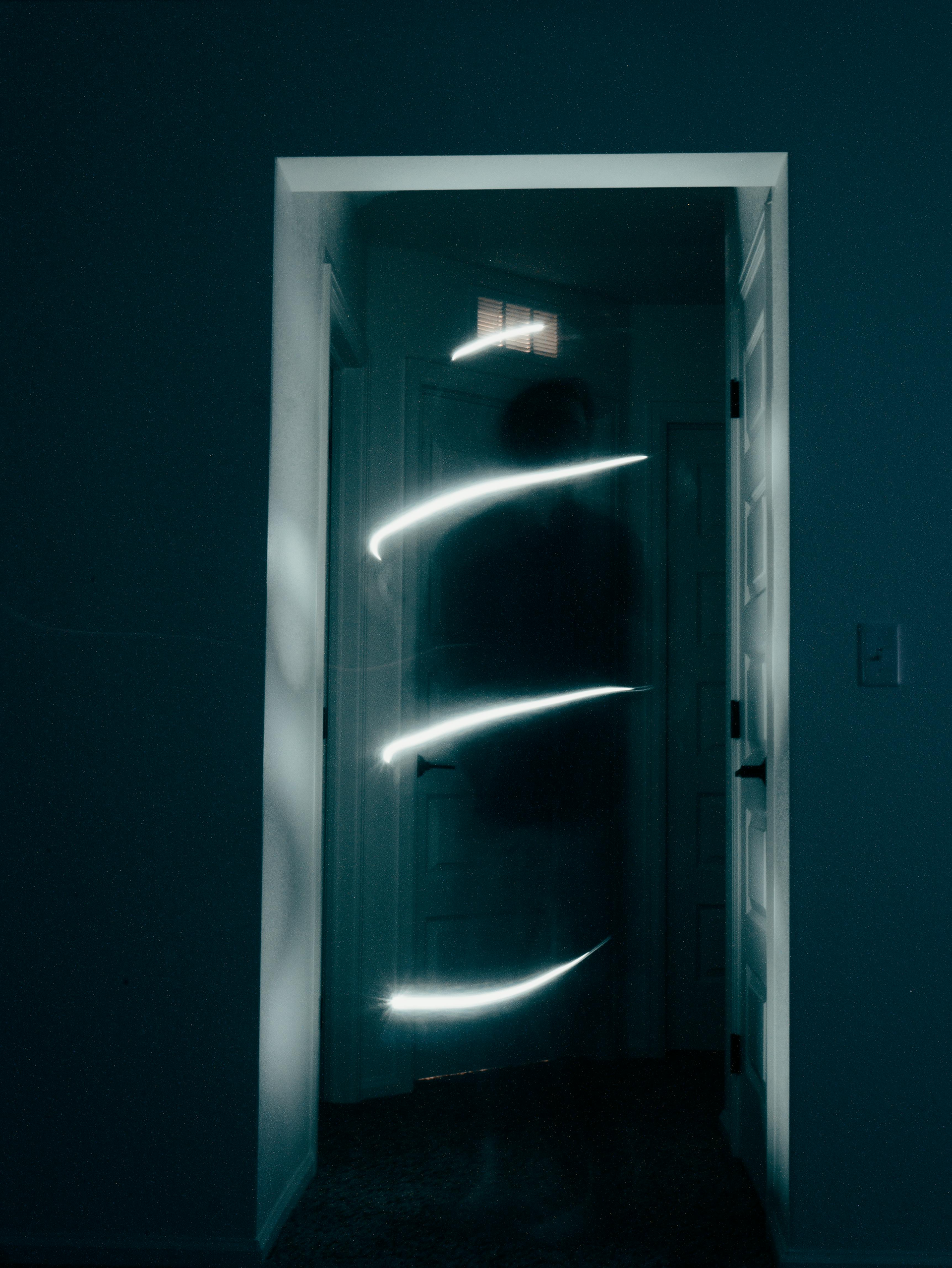 Dark room with an open doorway; white light streaks across the door as a figure moves.