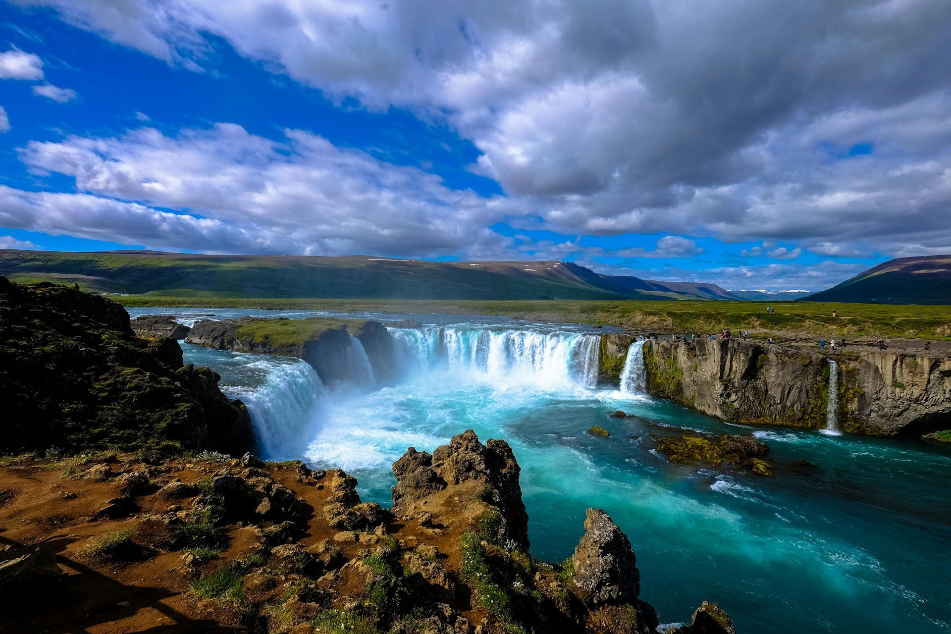 A wide, turquoise waterfall cascades over rocky cliffs under a partly cloudy, blue sky.