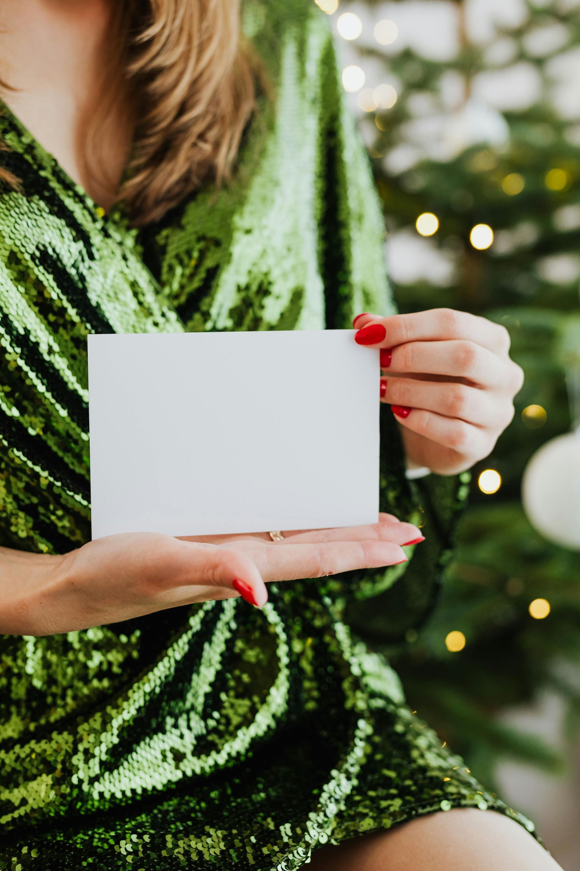 Woman in sparkly green dress holds a blank white card, red nails visible. Christmas tree in background.
