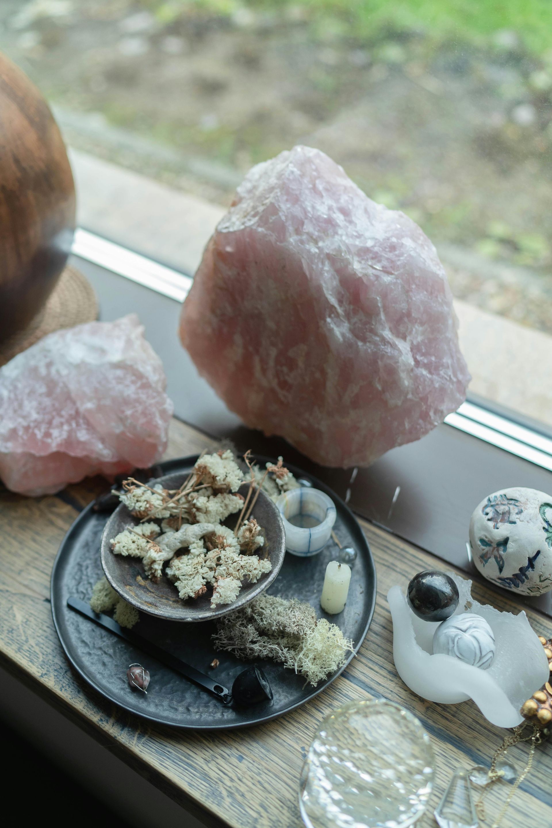 Pink rose quartz crystals on a windowsill, with a plate of dried flowers and other decorative objects.