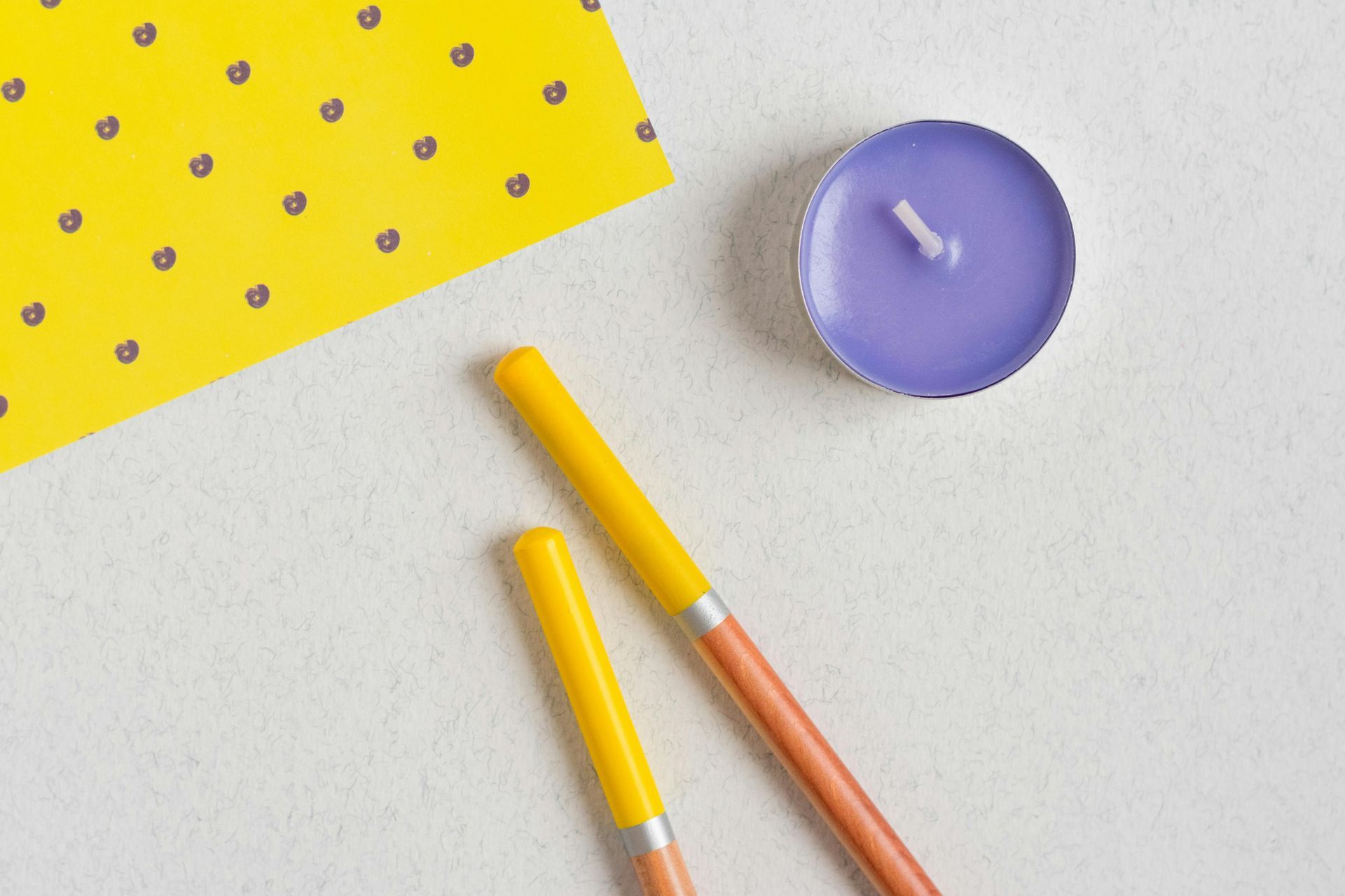 Yellow pencils, purple candle, and yellow card with black dots on a white surface.