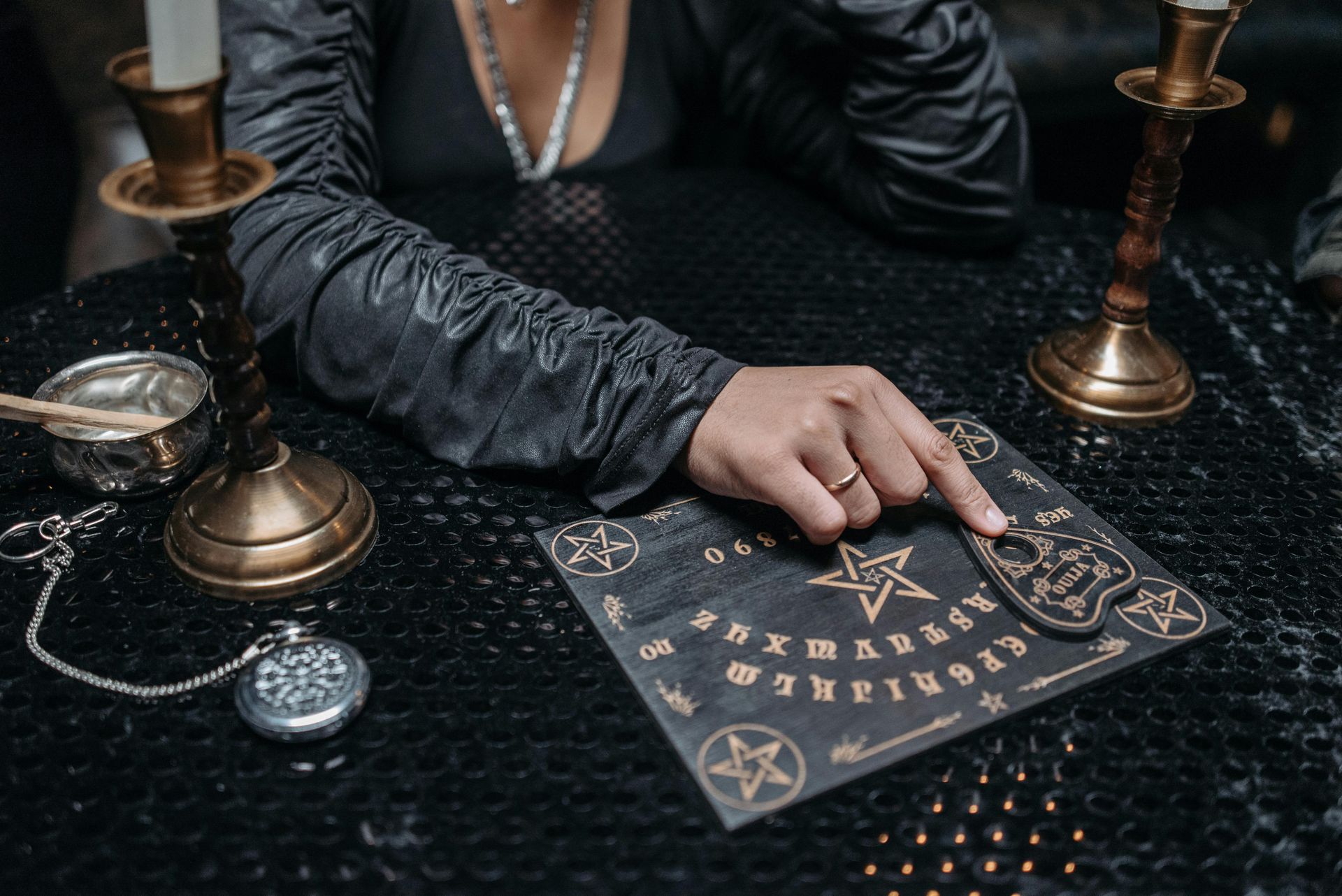 A person's hand on a Ouija board, surrounded by candles and a pocket watch, in a dimly lit room.