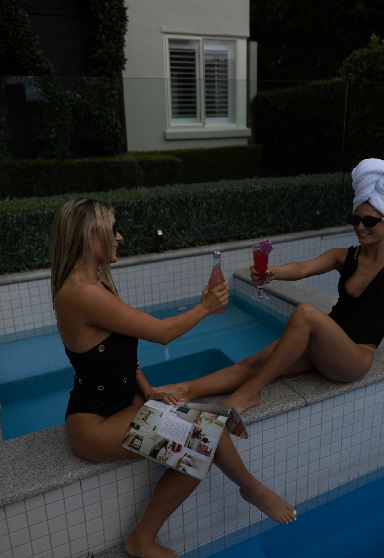 Two women are sitting on the edge of a swimming pool holding drinks.