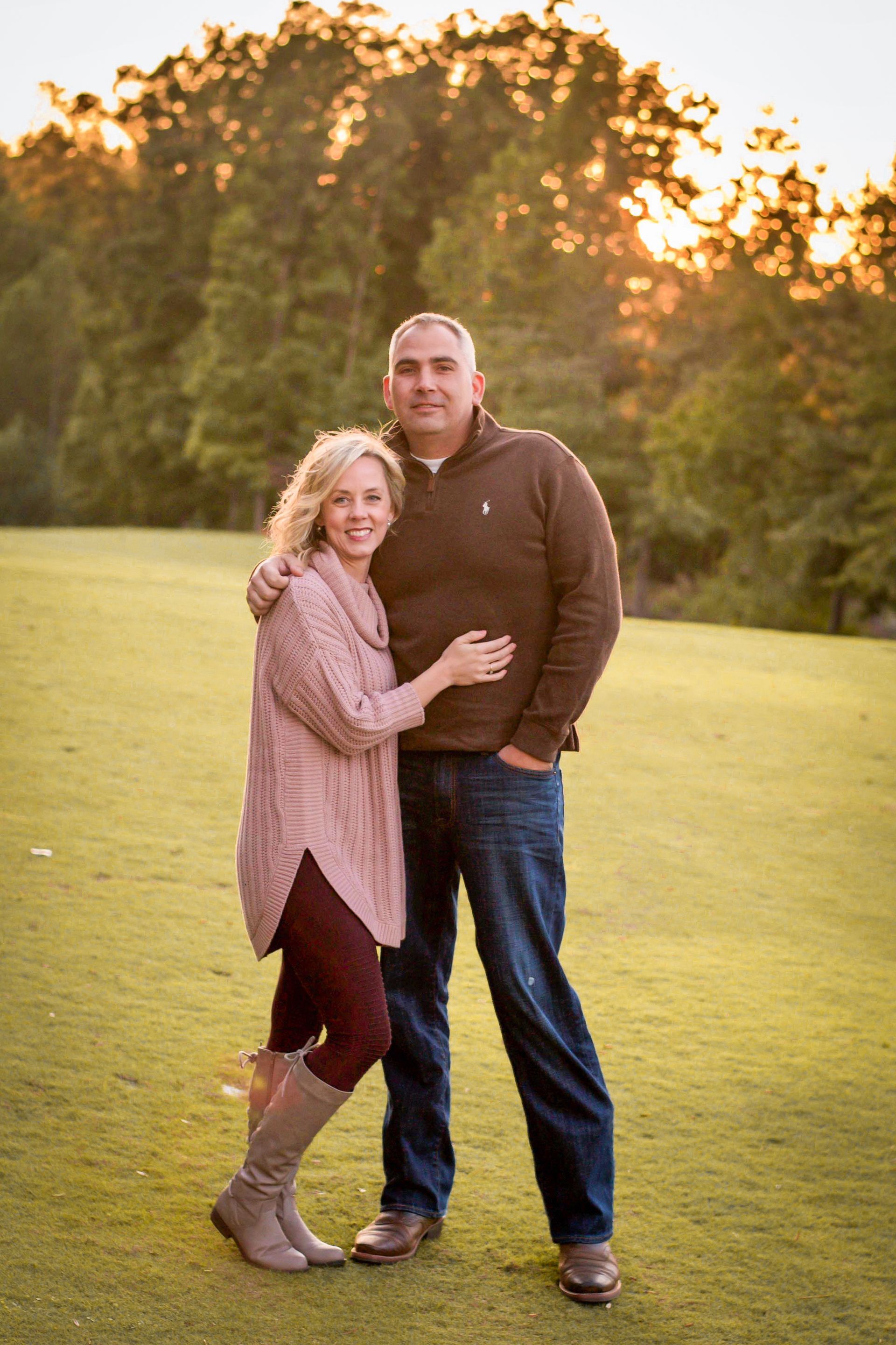 Couple embraces on a grassy field in front of a treeline, with a sunset in the background.