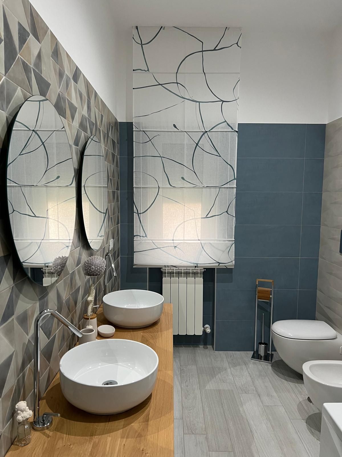 Bagno con piastrelle a motivi, due specchi rotondi, lavandini a appoggio e tenda a motivi.