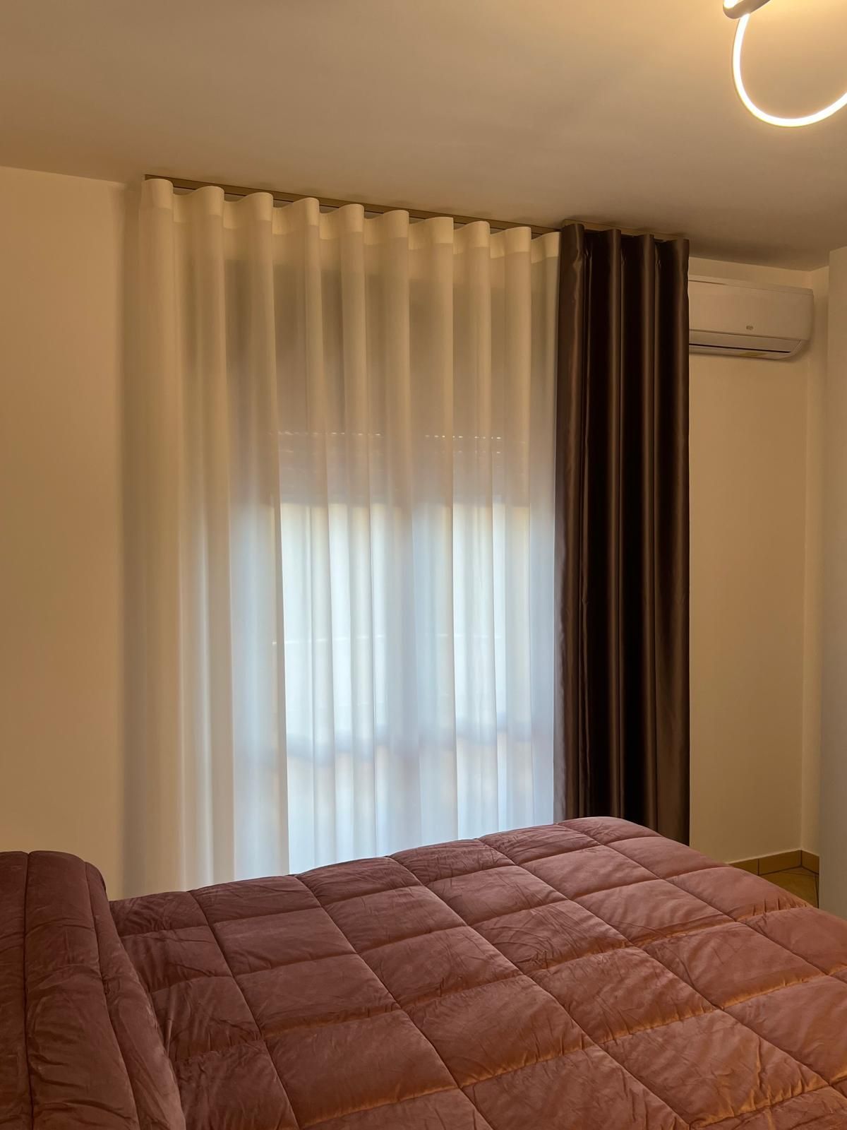 Letto con piumone rosa, finestra con tende bianche e marroni, aria condizionata sopra.