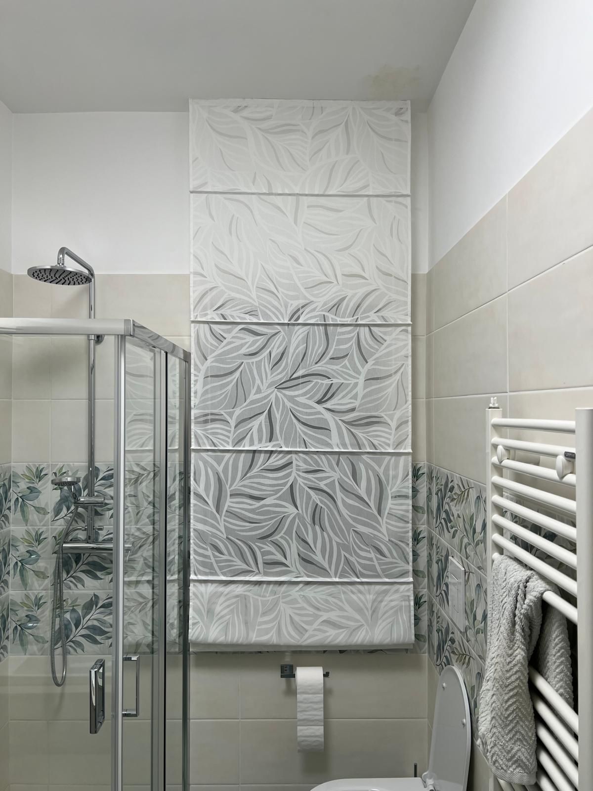 Bagno con doccia in vetro, piastrelle a fantasia e carta da parati con motivi a foglie. Toni del bianco e del grigio.