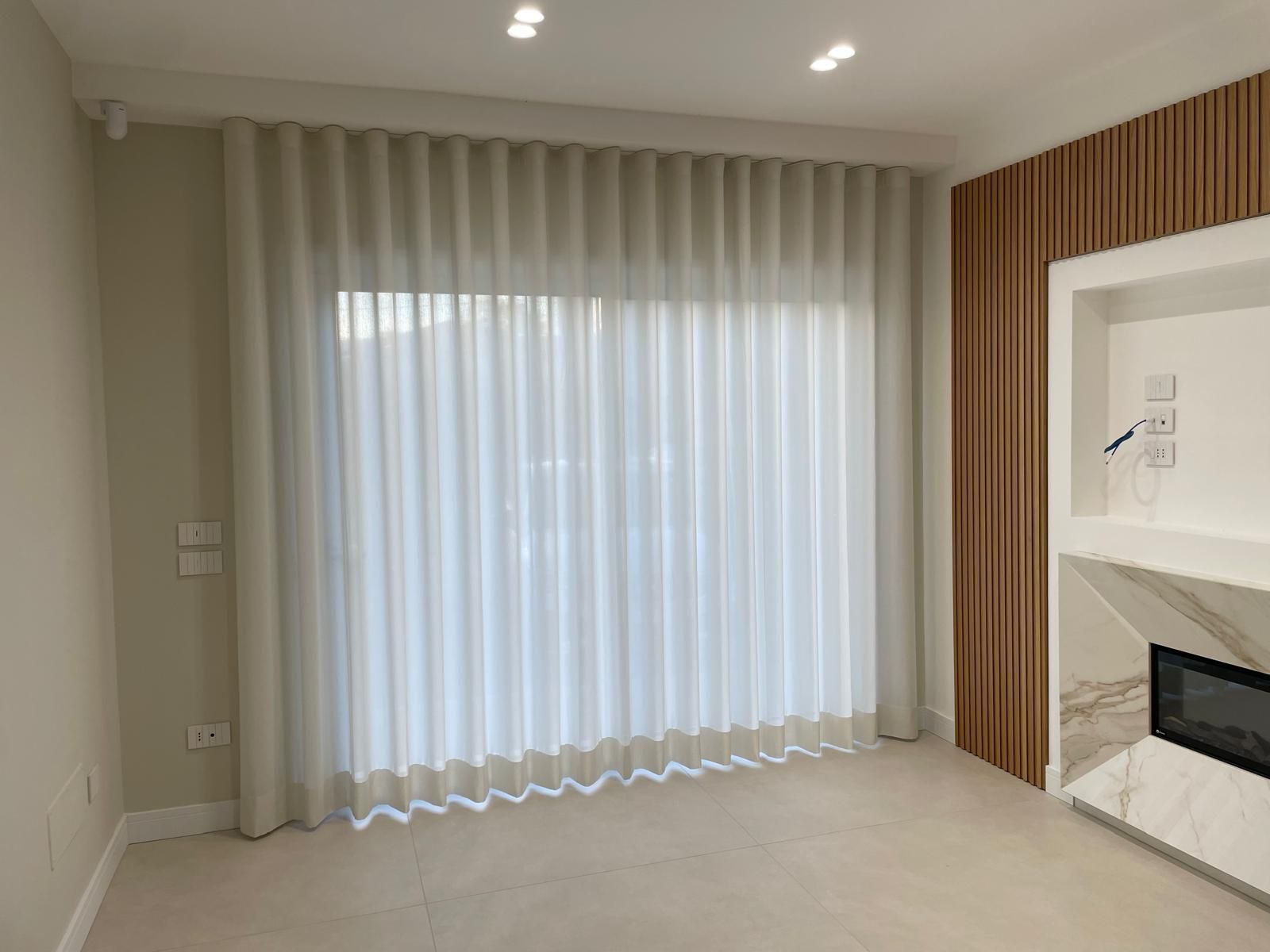 Tende plissettate beige coprono una grande finestra in una stanza moderna con pannelli in legno e camino.