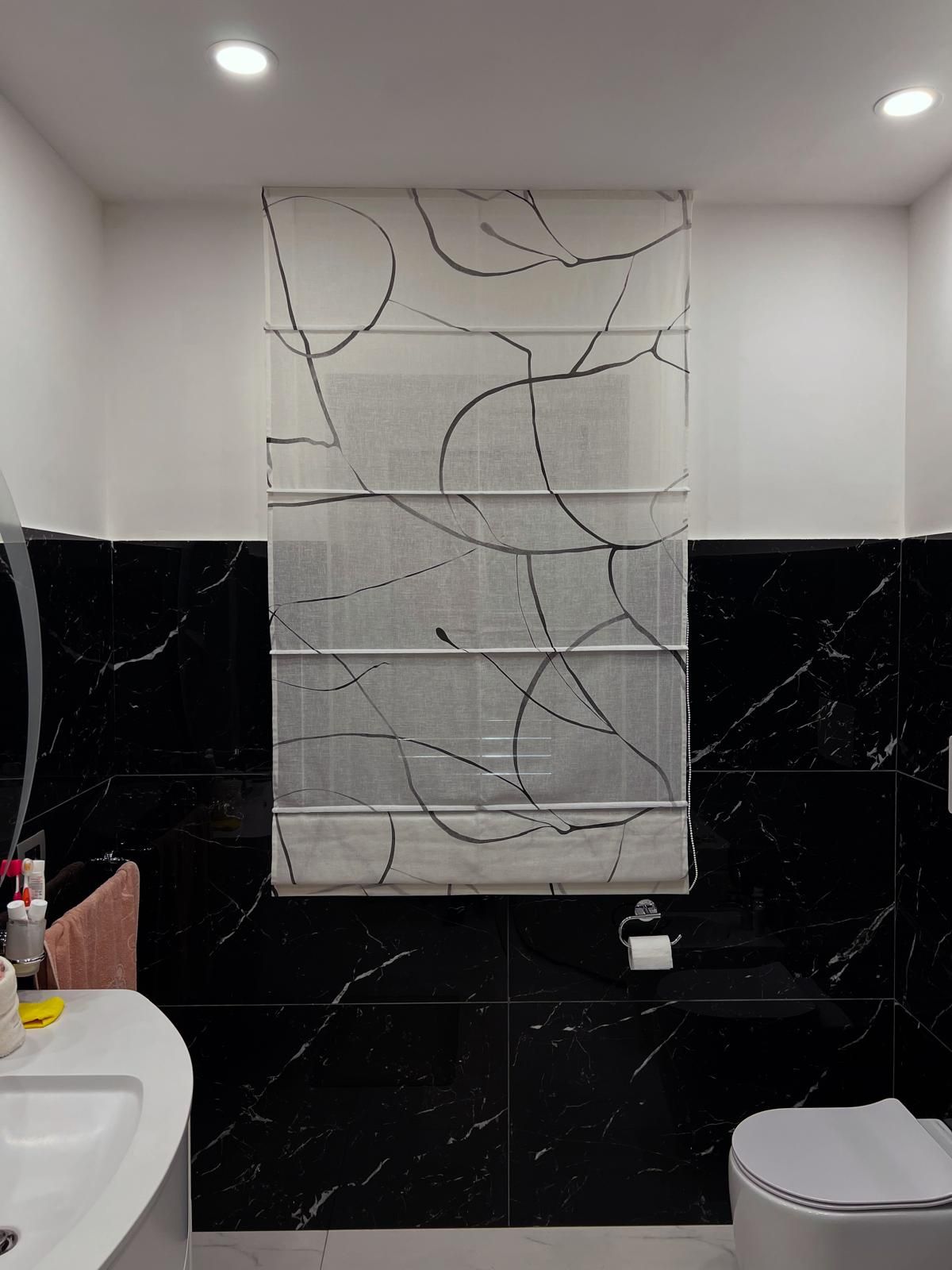 Bagno con piastrelle in marmo nero, lavandino e WC bianchi, finestra con tende a motivi astratti bianchi e neri.