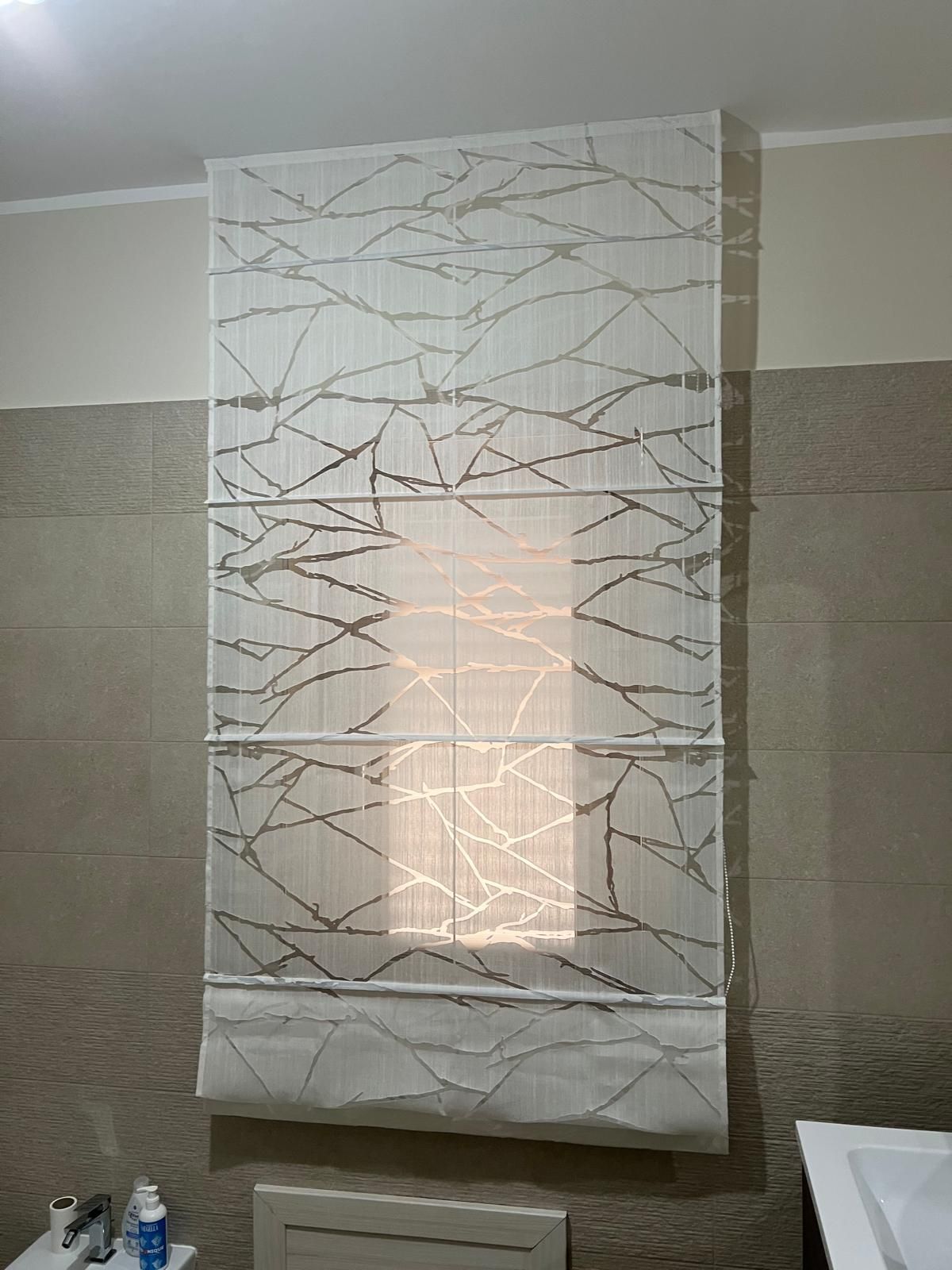 Tenda romana bianco sporco con linee grigie astratte sopra una piccola finestra in un bagno.