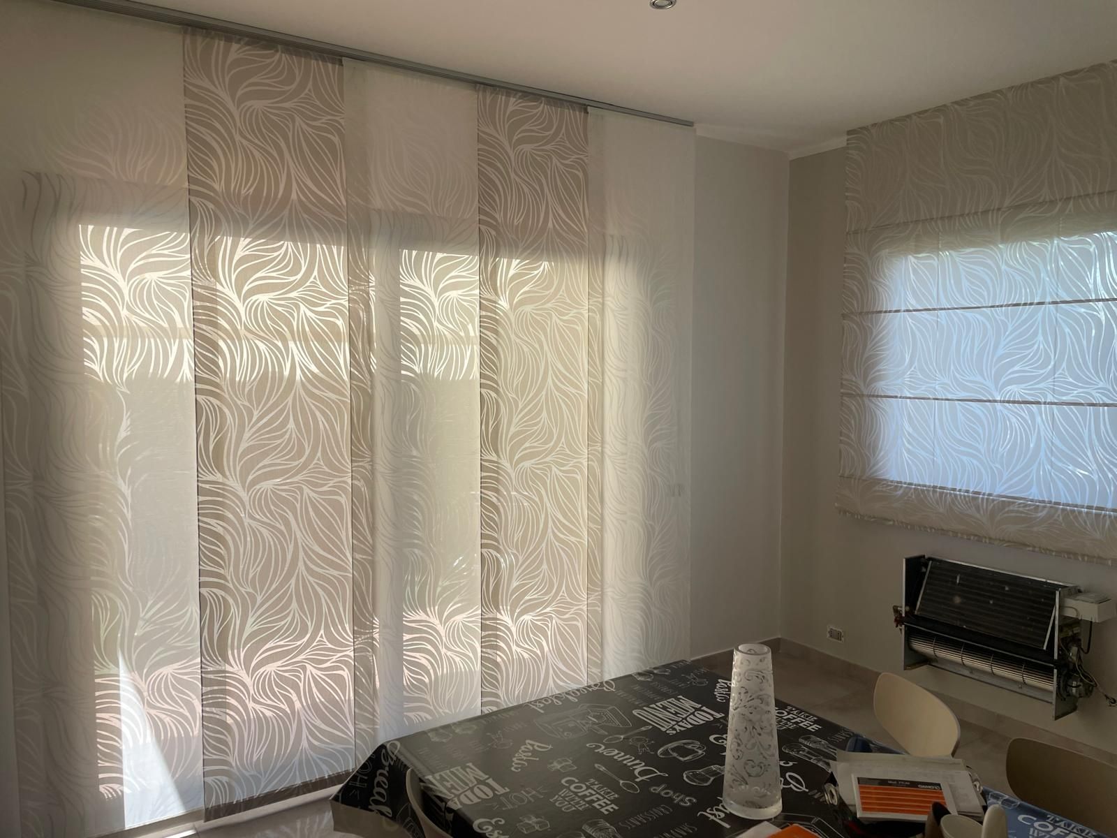 Tende a pannello e tenda romana con motivo a foglie beige; la luce del sole filtra attraverso le finestre.