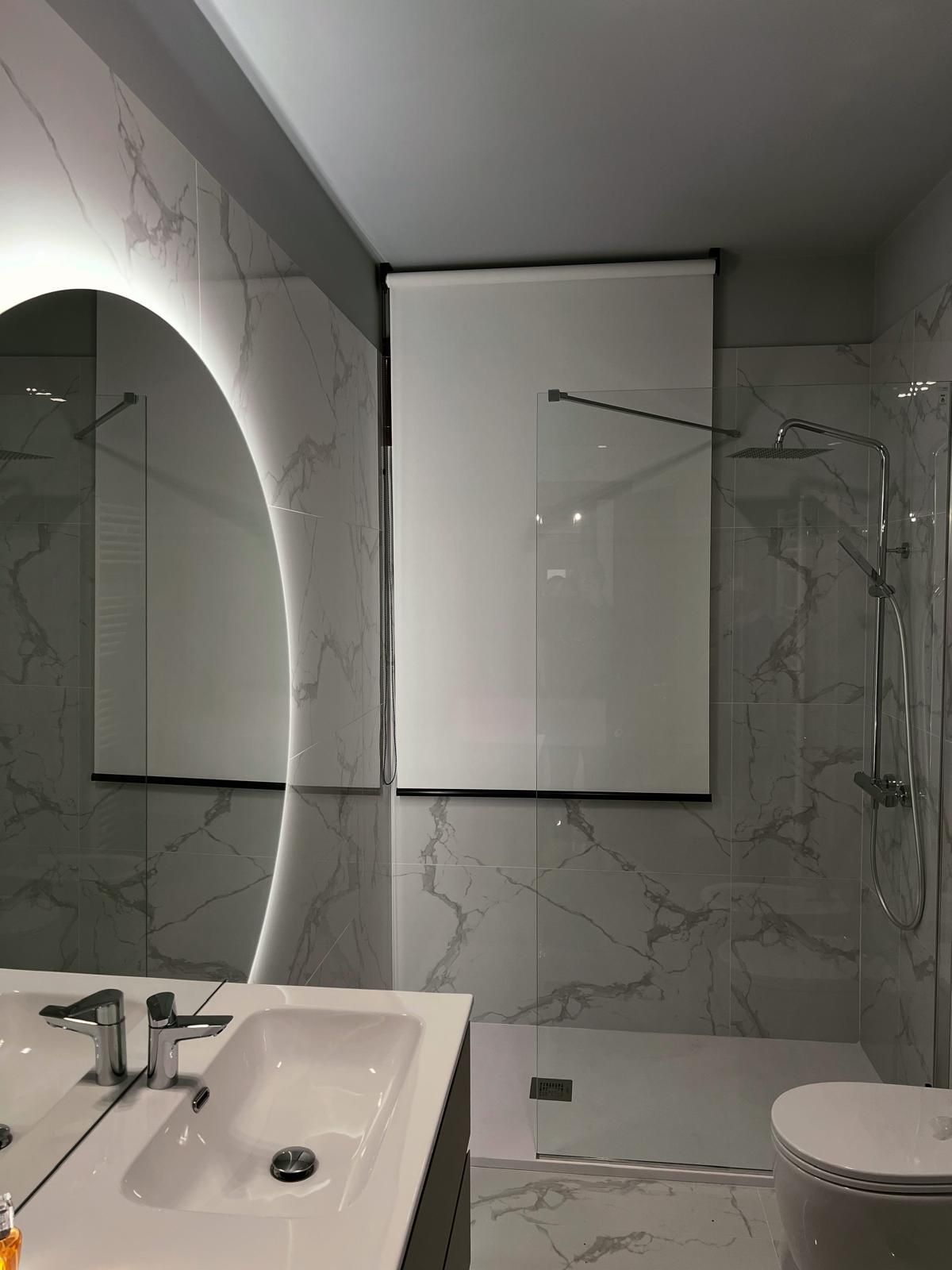 Bagno con piastrelle in marmo bianco, grande specchio e finestra con tenda bianca.