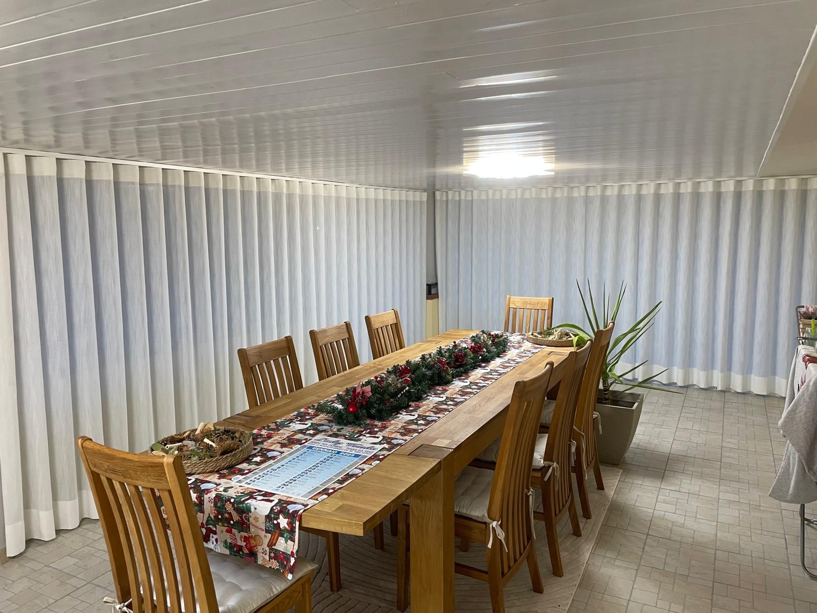 Zona pranzo con lungo tavolo in legno, dieci sedie, tende bianche e runner natalizio.