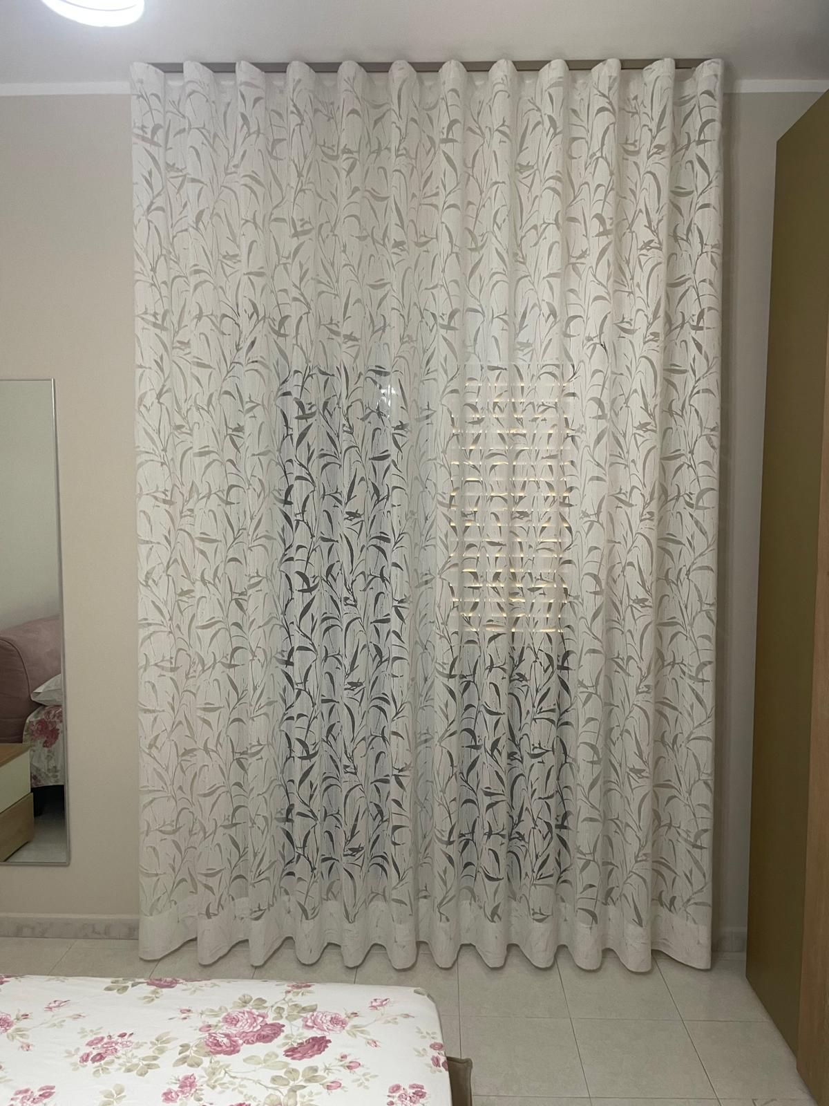 Tende bianche a fantasia appese davanti a una finestra. In primo piano si vede un letto.