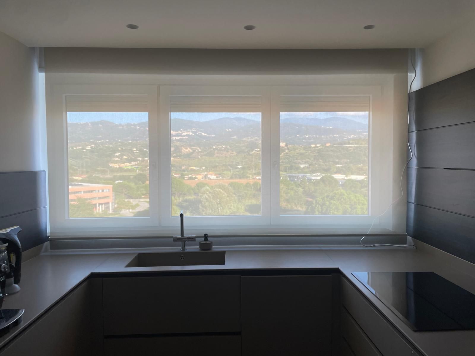 Cucina con tre finestre, vista sulla città, tende dai toni neutri, piano di lavoro, lavello e piano cottura.