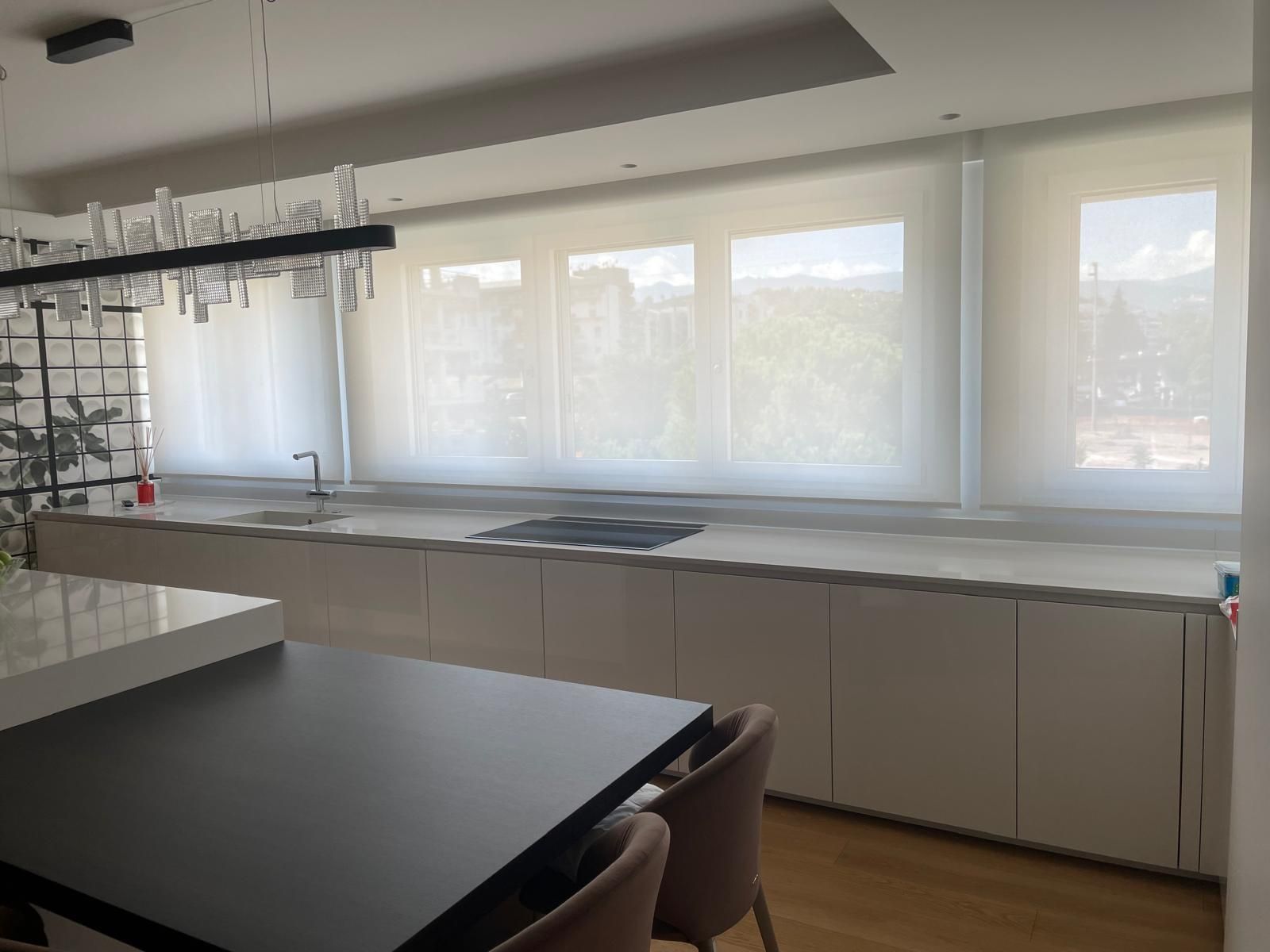 Cucina moderna con lungo piano di lavoro, mobili bianchi e tende trasparenti alle finestre.