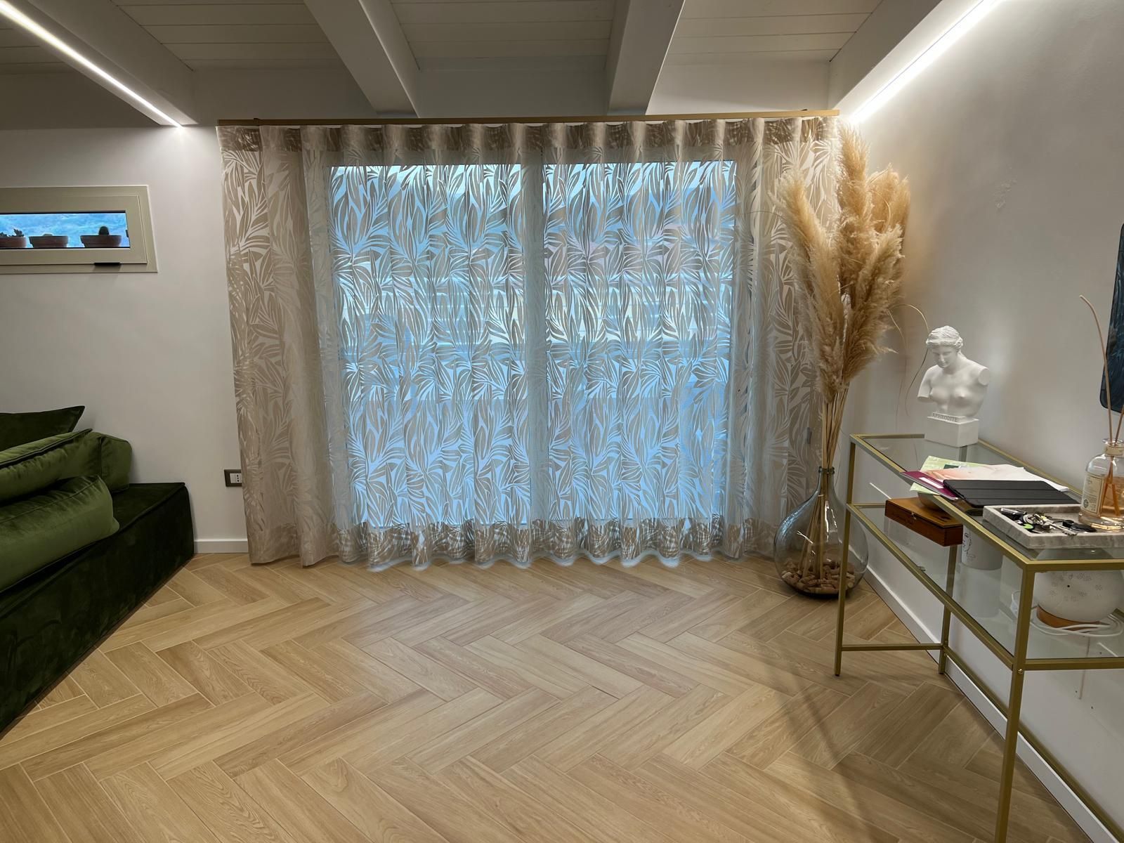 Soggiorno con tende dai colori chiari, pavimento in legno a spina di pesce e consolle con bordi dorati e erba della pampa.