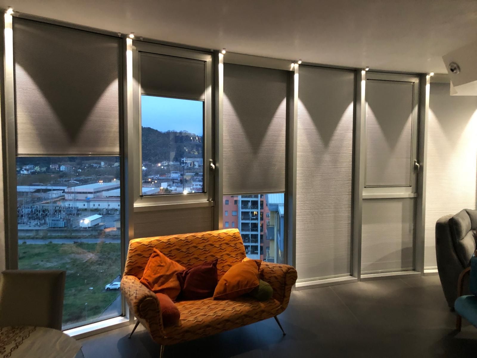 Soggiorno con finestre curve e persiane, con vista sul paesaggio urbano. Sotto, una poltrona decorata.
