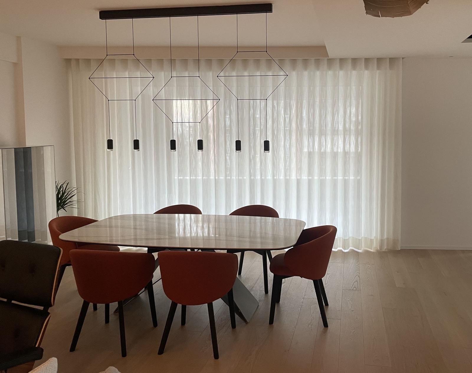 Sala da pranzo con tavolo con piano in marmo e sedie arancioni, sotto un lampadario geometrico e tende trasparenti.