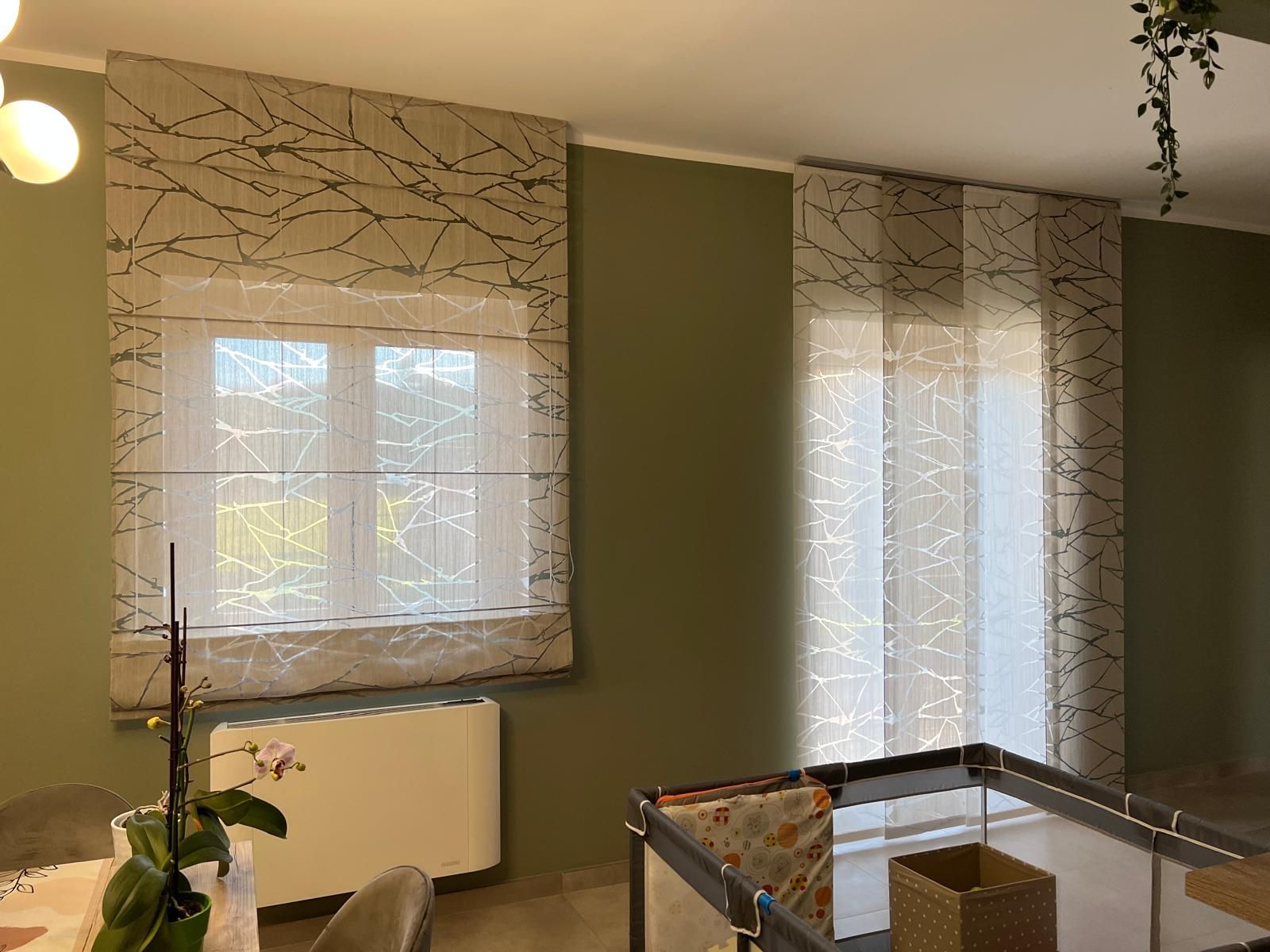 Tende romane e tende a pannello davanti a una finestra, beige con motivi a linee nere, contro una parete verde.