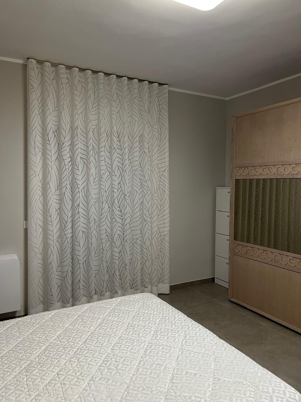 Camera da letto con tende bianche a fantasia, armadio in legno chiaro e letto con copriletto trapuntato bianco.