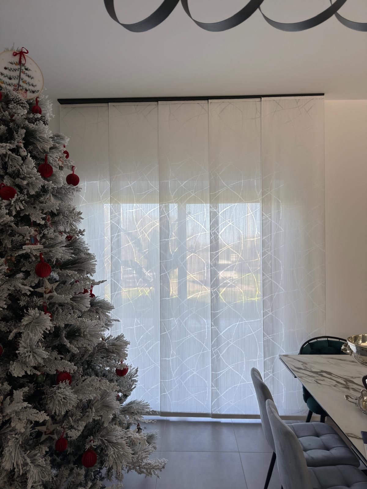 Albero di Natale accanto a tende bianche trasparenti in una stanza ben illuminata. Tavolo da pranzo con sedie sulla destra.