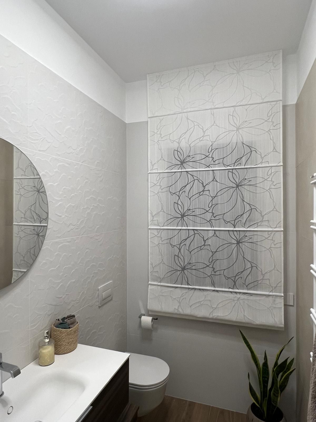 Bagno con tenda romana decorata sopra la finestra, uno specchio rotondo e una piccola pianta.