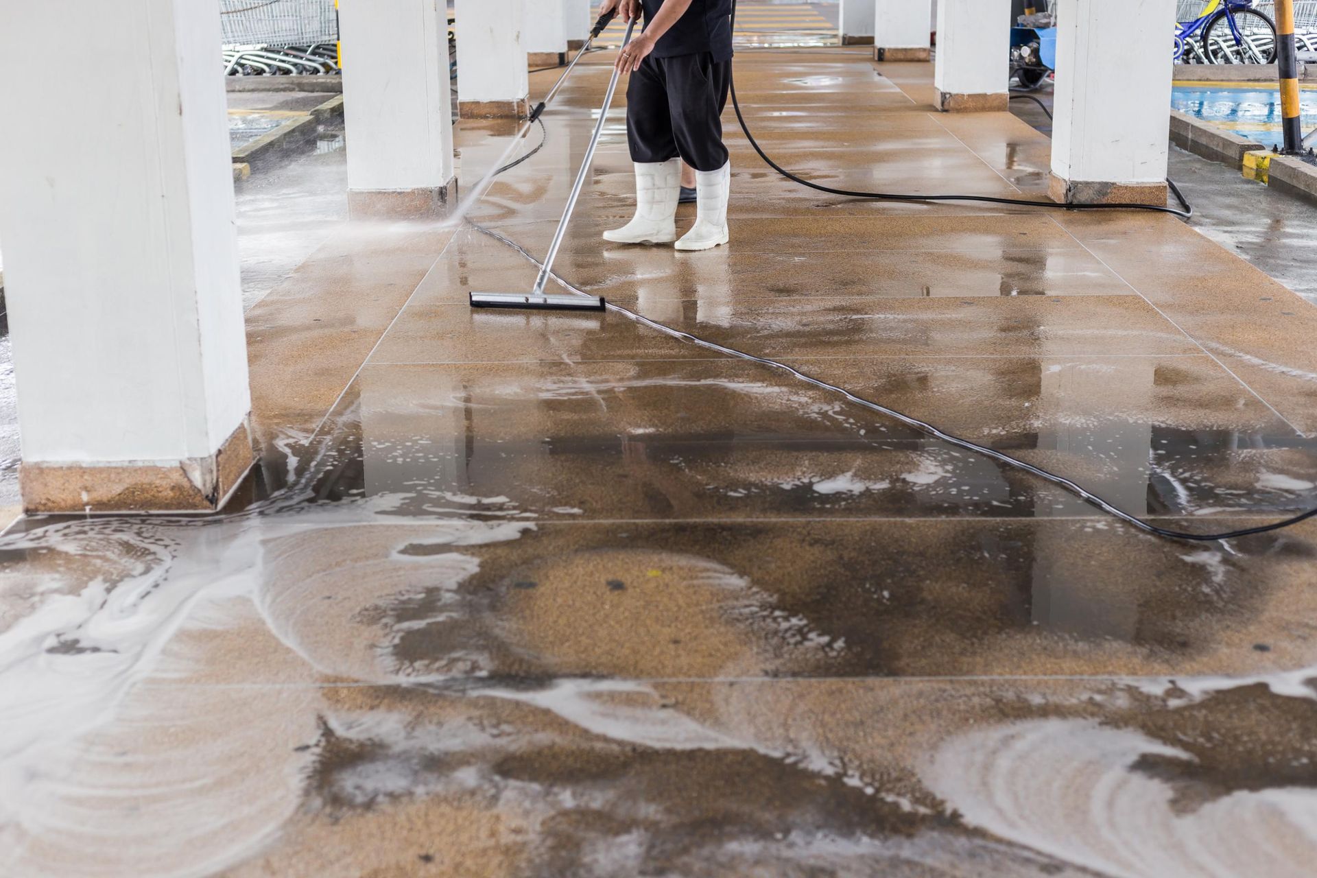 Person power washing a concrete floor with water and soap near white pillars.