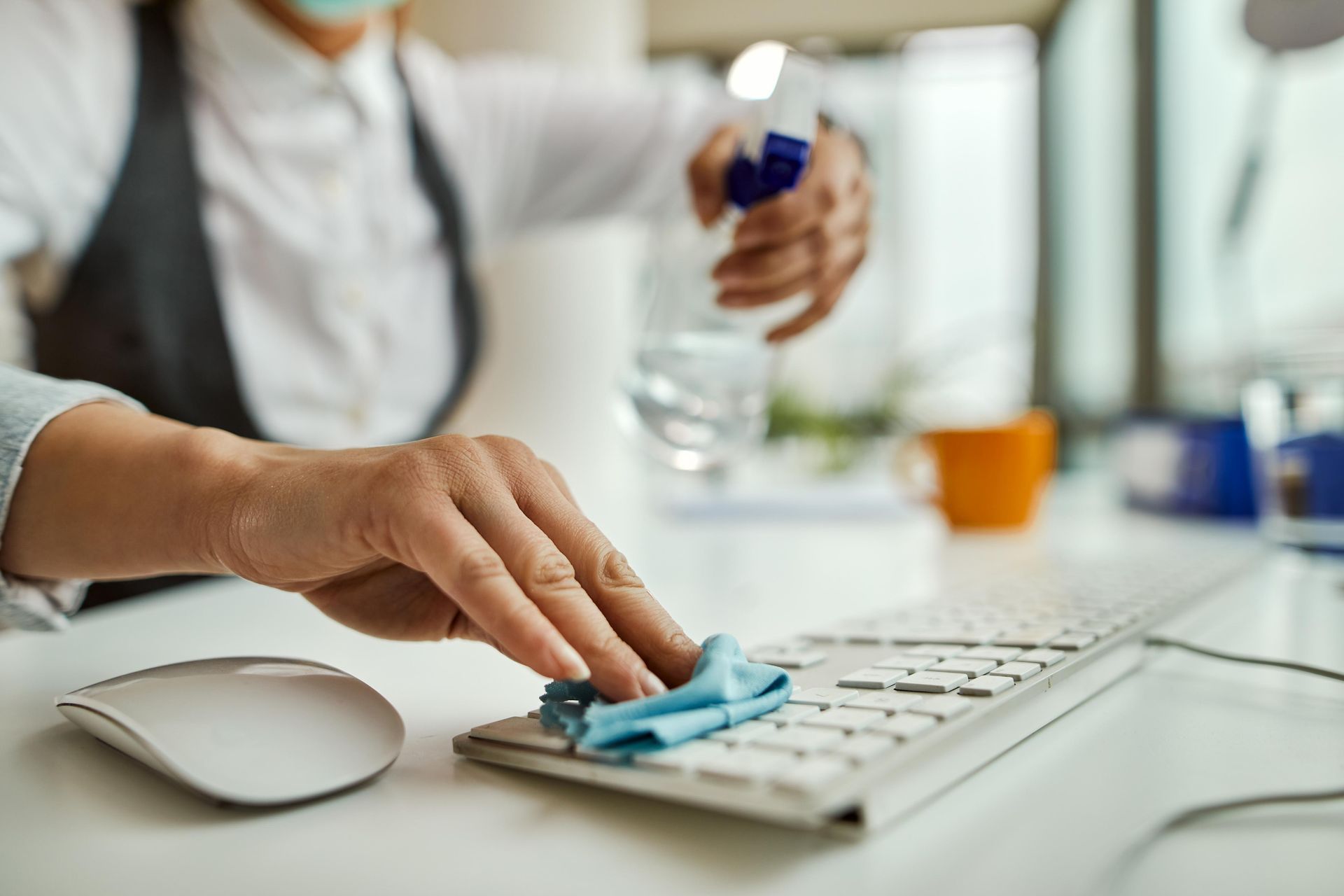 Person sanitizes a keyboard with a spray bottle and cloth at a desk.
