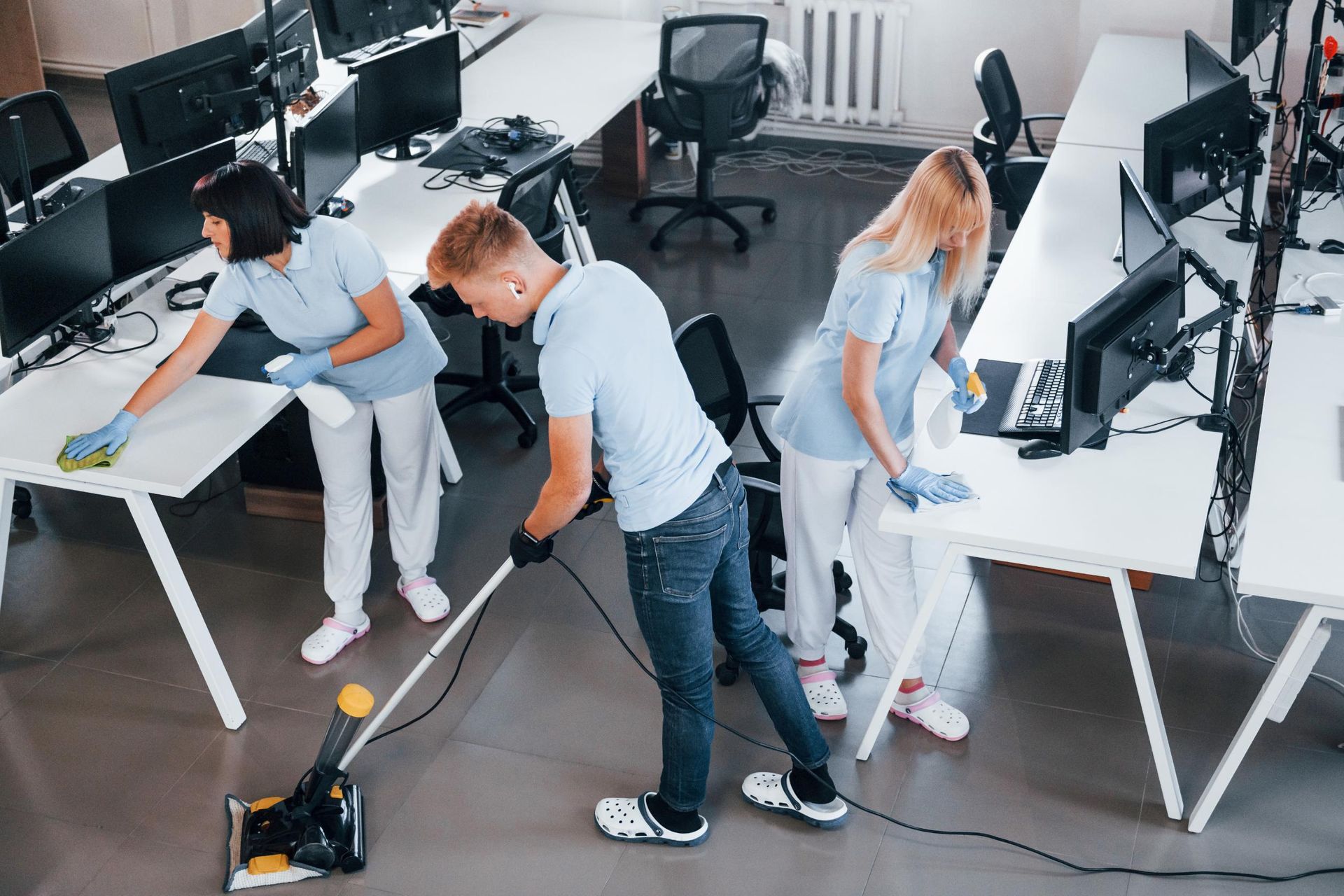 Three people cleaning an office. One vacuums while the others wipe down desks. Bright, modern office space.