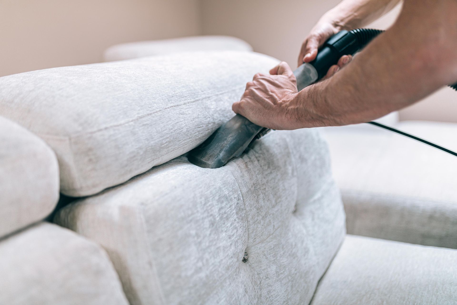 Person vacuums a light-colored couch, nozzle cleaning between cushions.