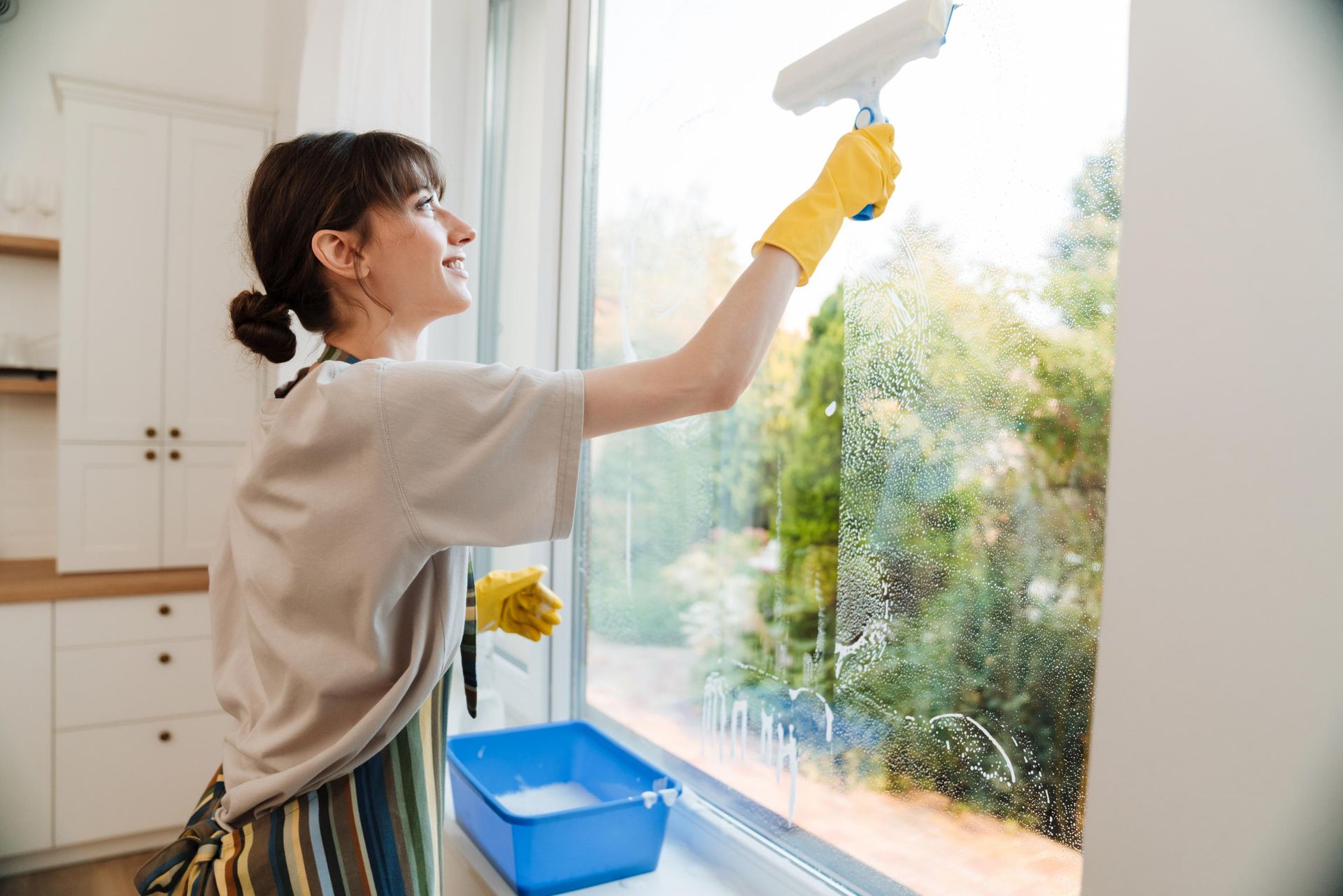 Woman wearing yellow gloves cleaning a window with a squeegee. She stands inside a home, looking outside.