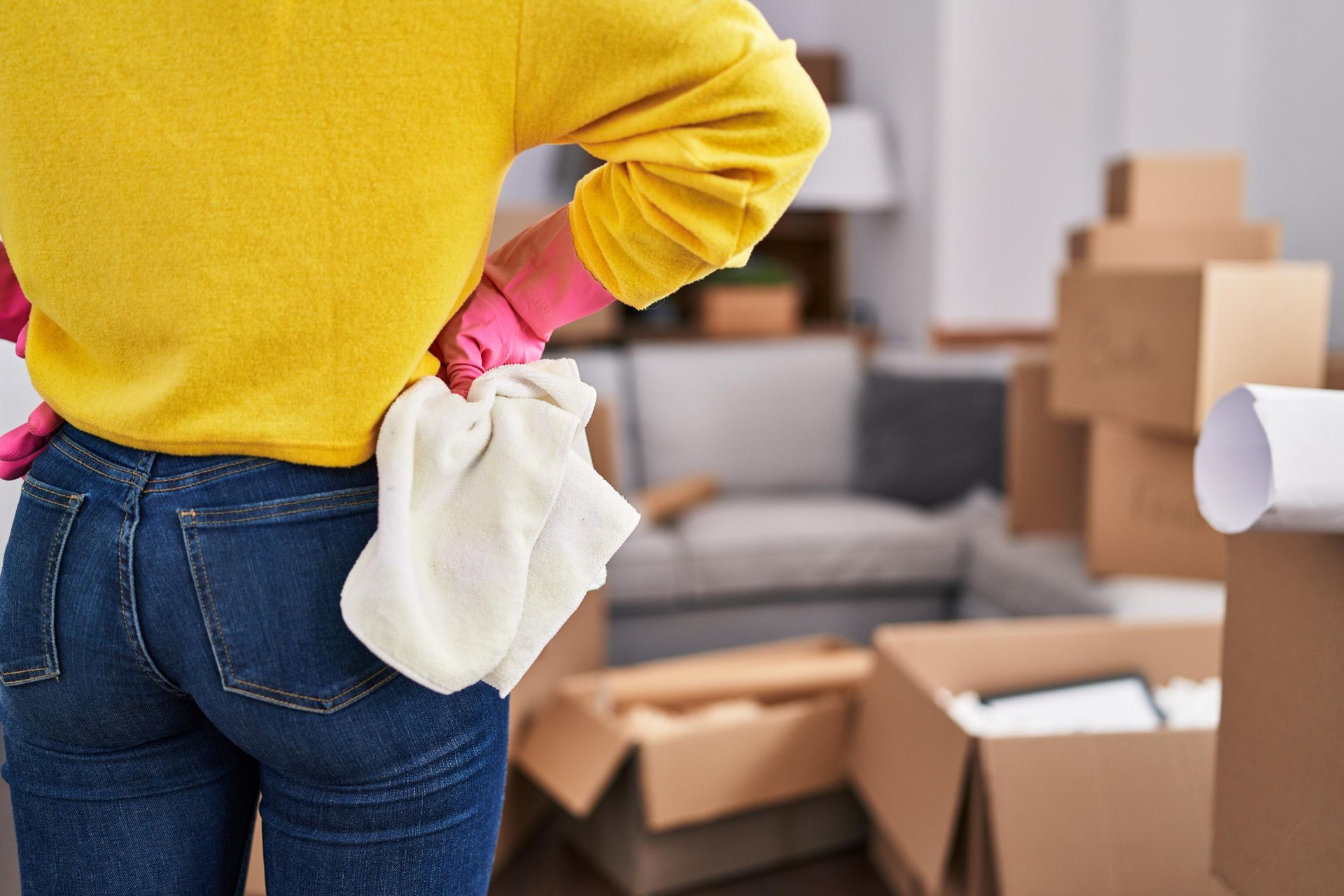 Woman in pink gloves and yellow sweater, jeans, stands with hands on hips in a room with moving boxes.