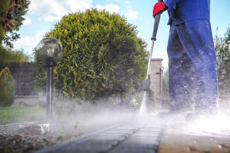 Person pressure-washing a paved area beside a hedge, with water spraying across the ground