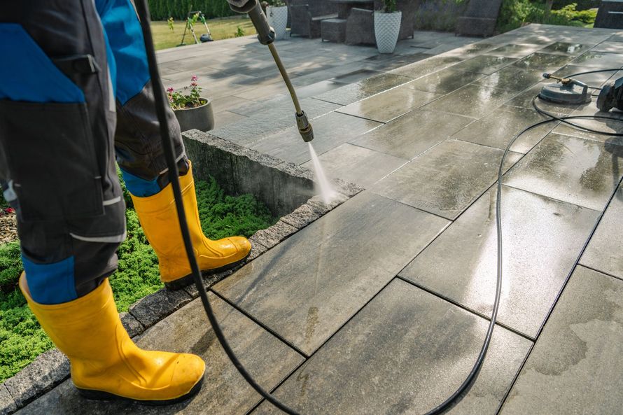 Person pressure-washing a patio, wearing yellow boots, with water spraying across wet stone tiles.
