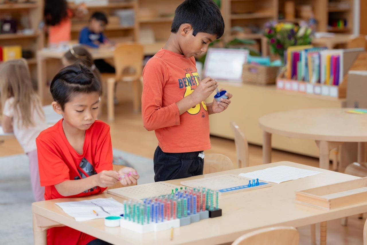 Two Montessori students are sitting at a table in a classroom working with math material.