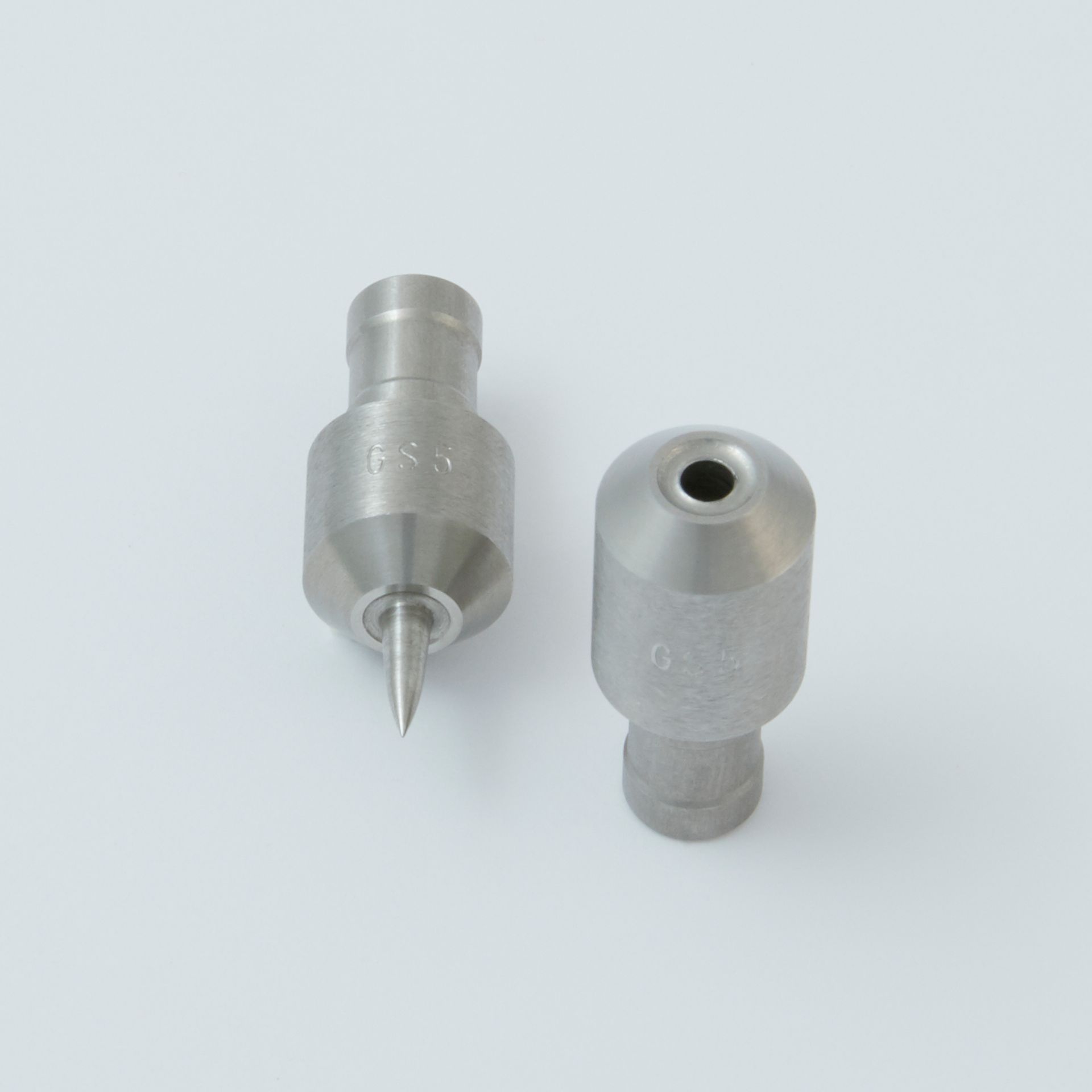 Two metallic nozzle components on a white background: one with a pointed needle tip and the other with an open circular port.