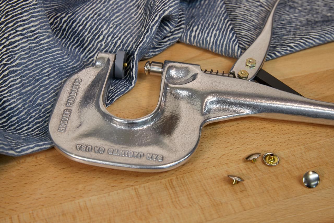Silver grommet tool on a wooden surface, with blue fabric and grommets. Scissors lie to the side.