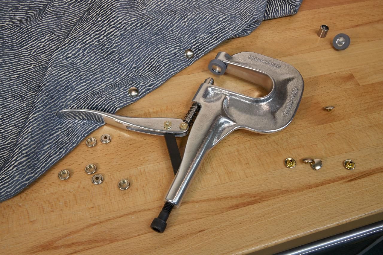 A silver rivet press tool, fabric, and rivets on a wooden surface.