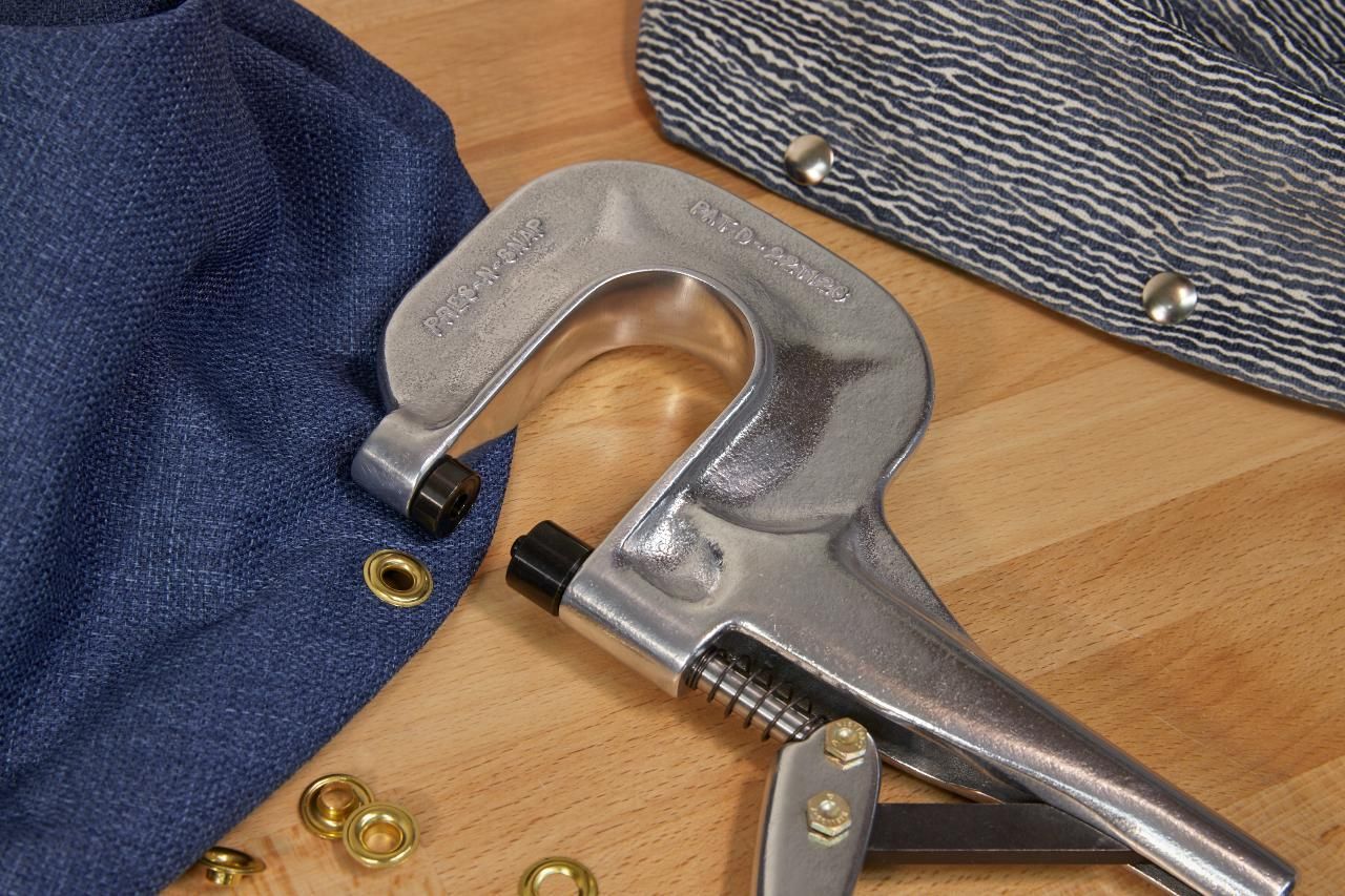 Eyelet press tool with blue fabric, gold eyelets, and a gray fabric bag.