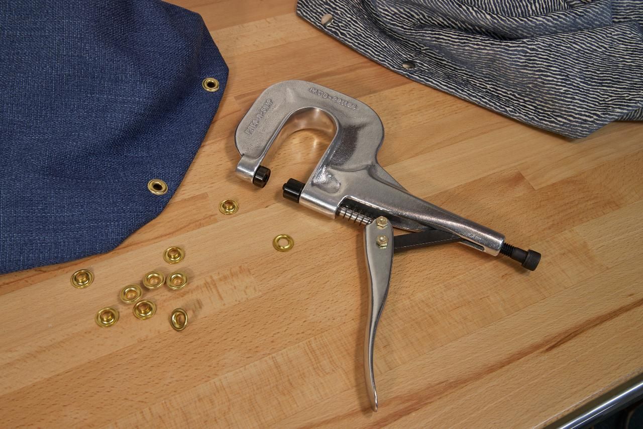 Metal eyelet tool with gold eyelets on wooden surface, with blue fabric and another fabric in the background.