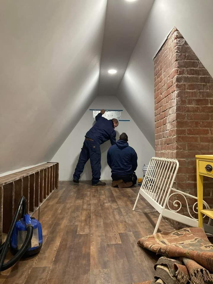 Two men are working on a wall in an attic.
