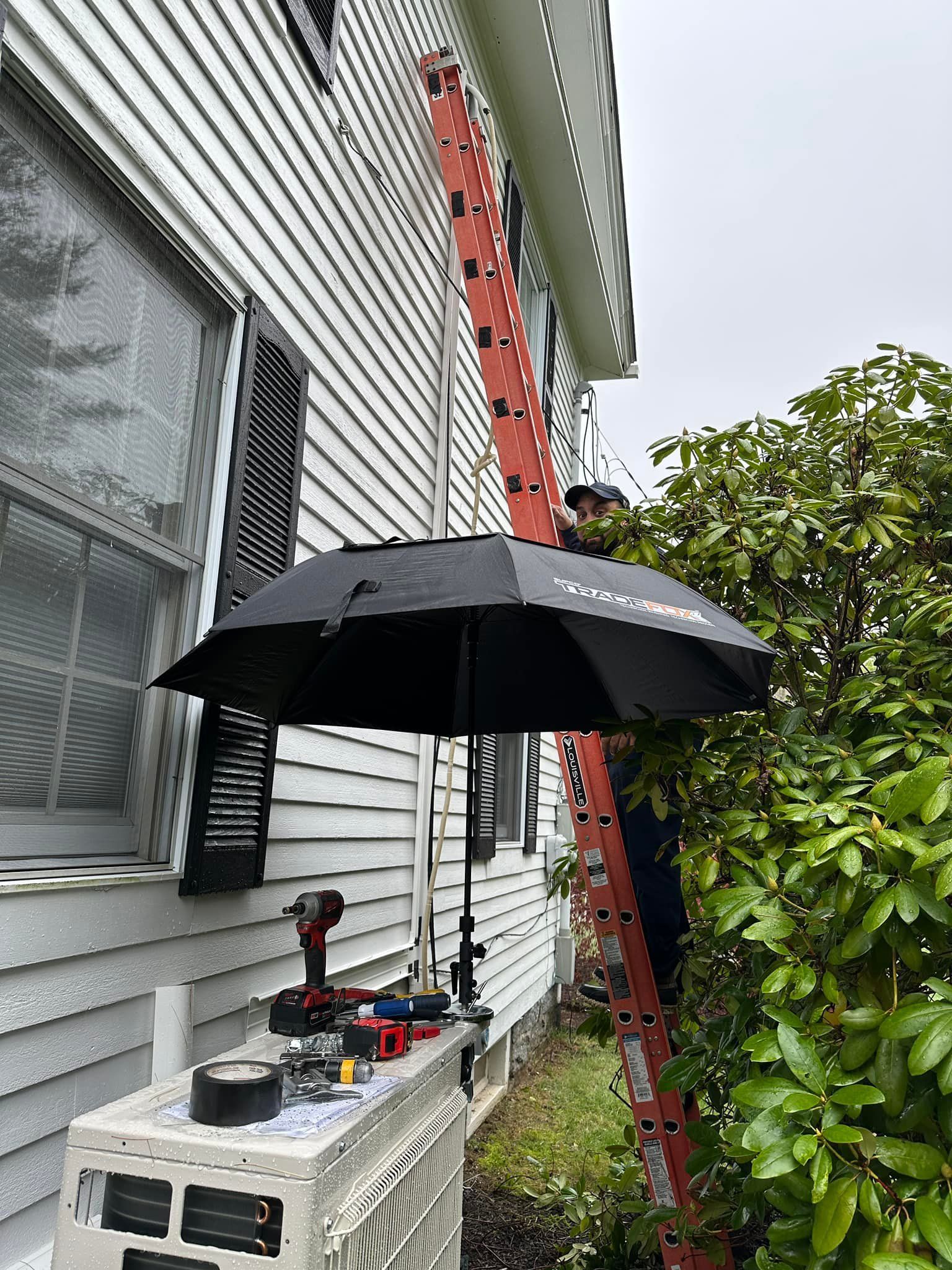 A man is standing on a ladder next to a house with an umbrella.