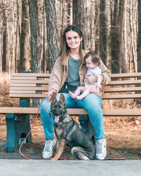 Woman sitting on park bench, holding a baby, with a German Shepherd dog. Outdoor setting with trees.